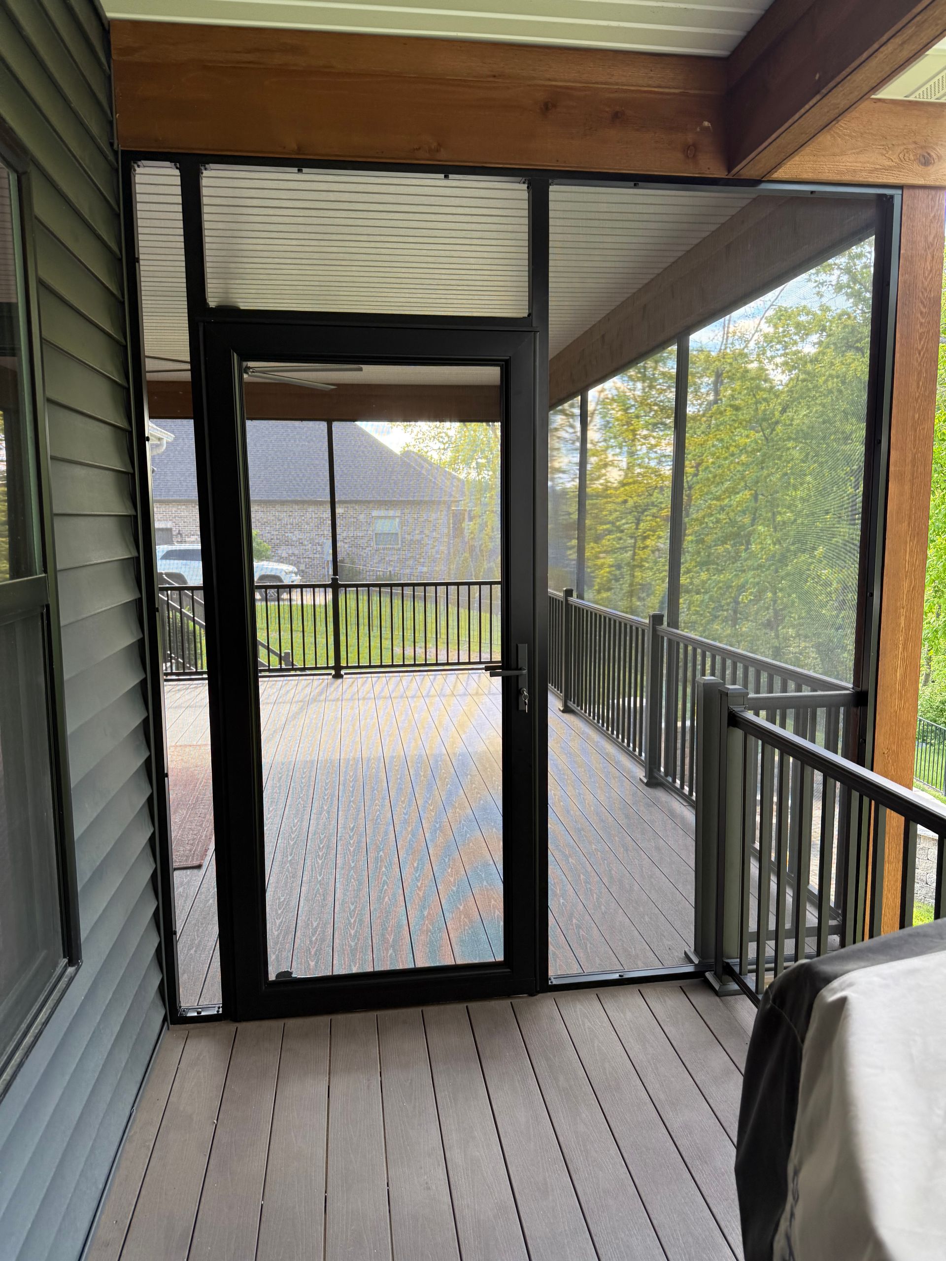 Screened-in porch with black door, gray deck, and wooden railing. Exterior view of house and trees beyond.