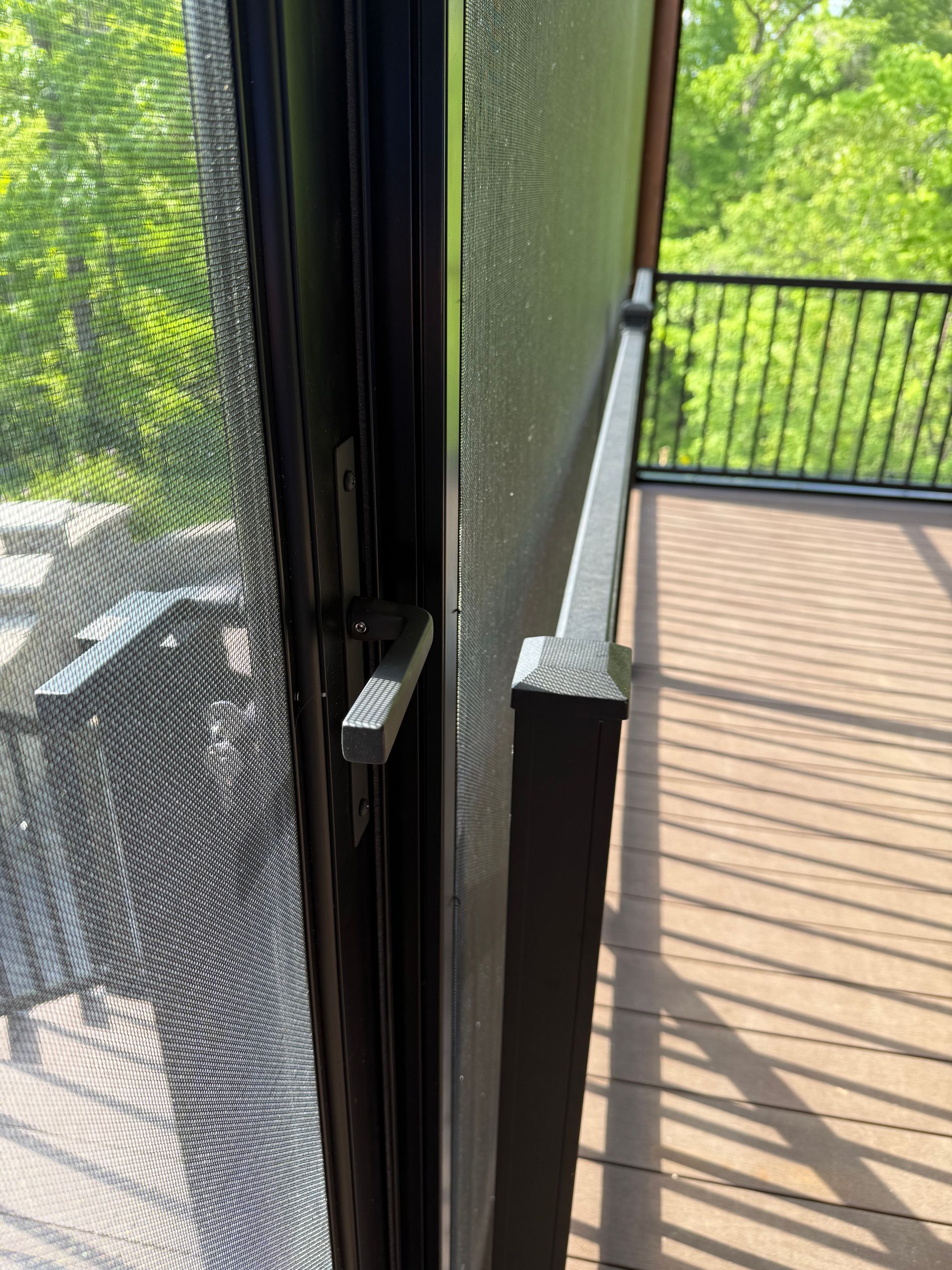 Black screen door ajar, handle visible, leading to a deck with black railing, overlooking a wooded area.