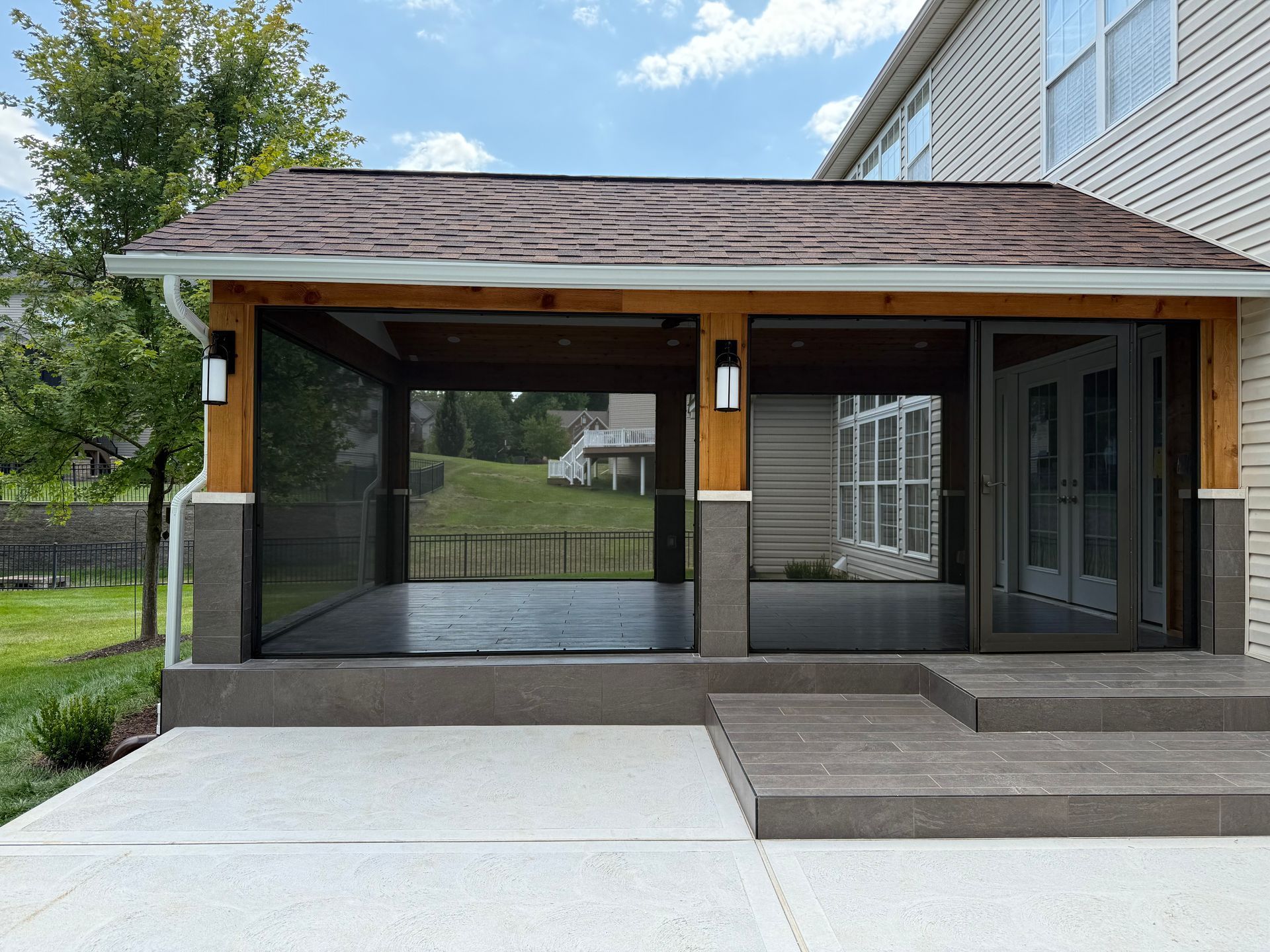 Screened-in patio extension with brown wood frame, gray steps, and grass lawn viewed from a gray concrete patio.
