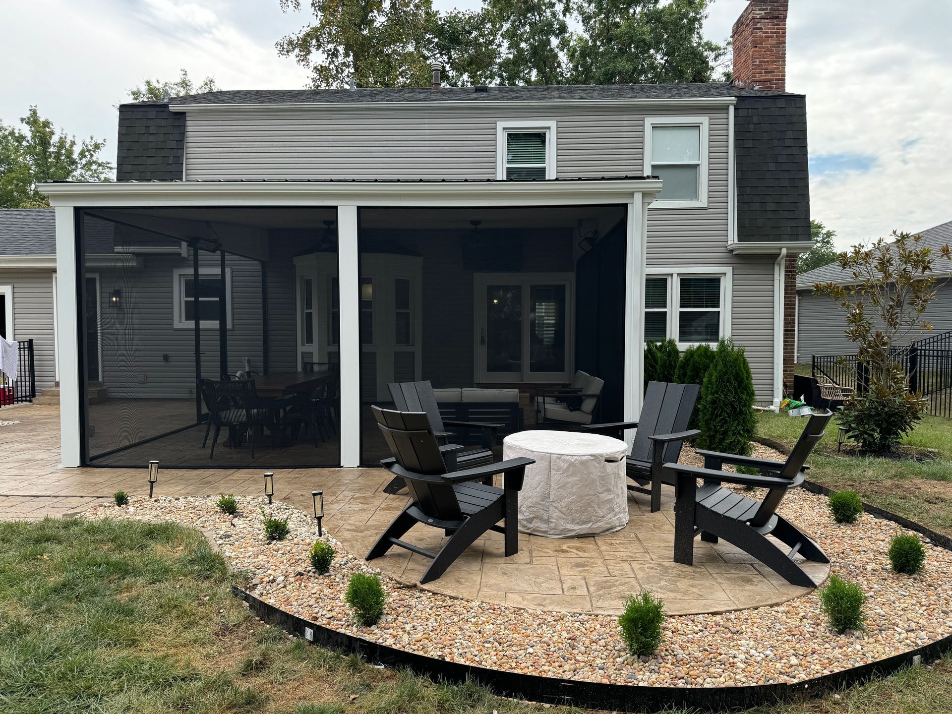 Screened-in patio with seating around a fire pit, bordered by tan gravel and small green bushes; gray house in the background.