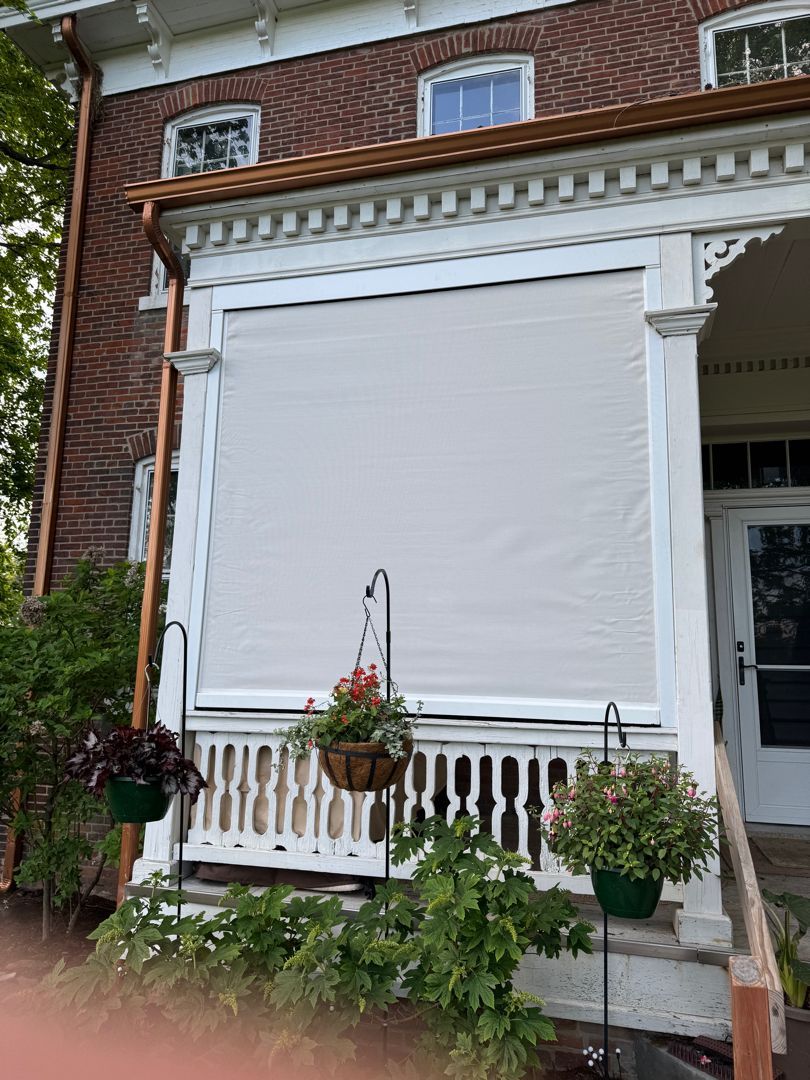 Beige sun shade on a porch with hanging flower baskets and white railing. Brick building with copper gutters.