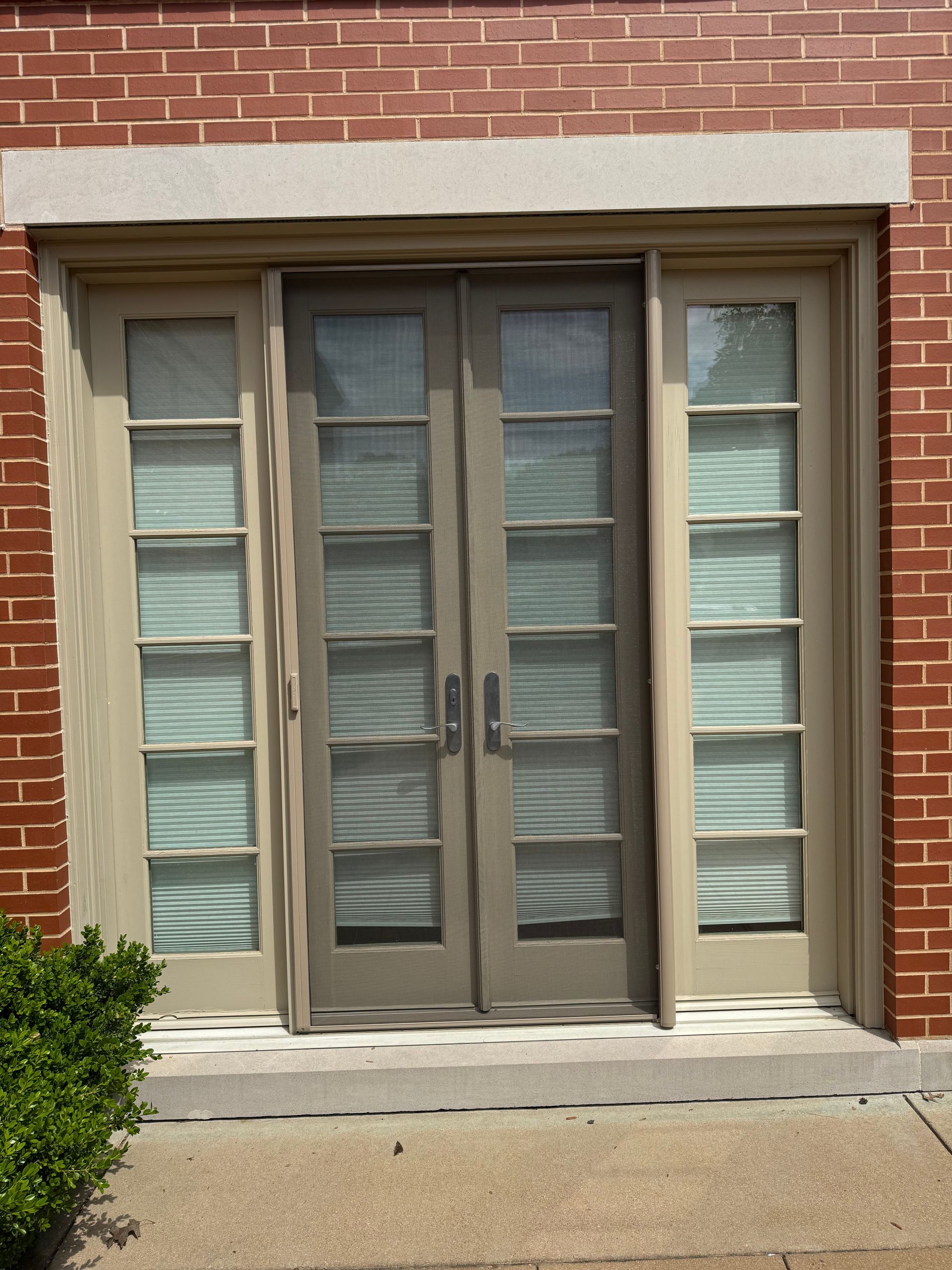 Exterior view of a building with light beige double doors and side windows with blinds, framed by red brick.