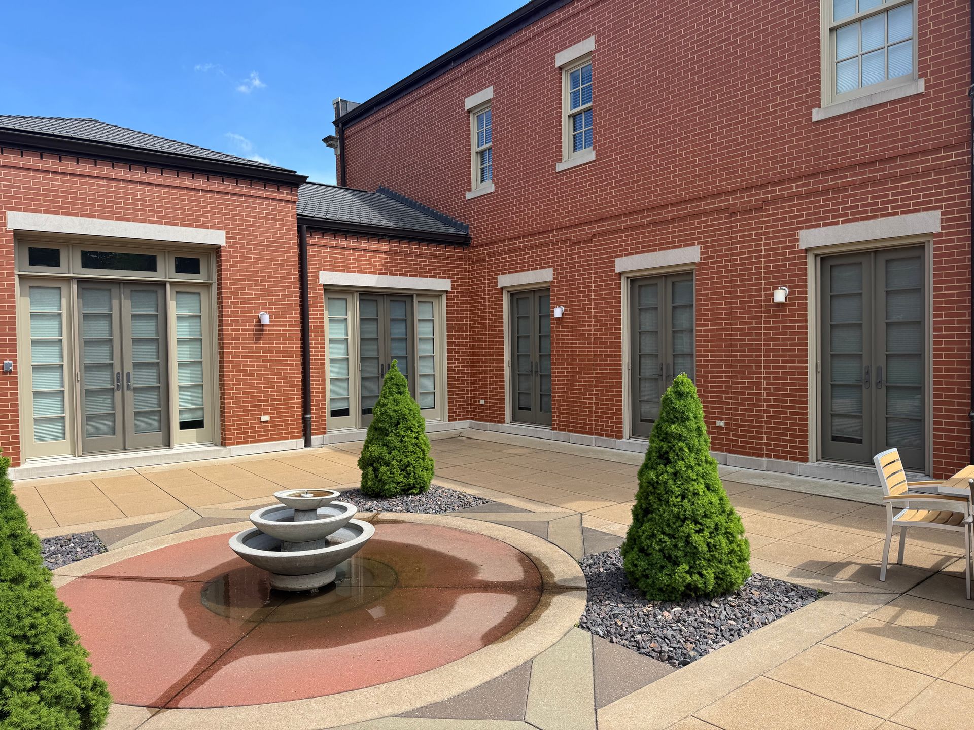 Brick courtyard with fountain, trimmed trees, and gray doors against a red brick building under a blue sky.