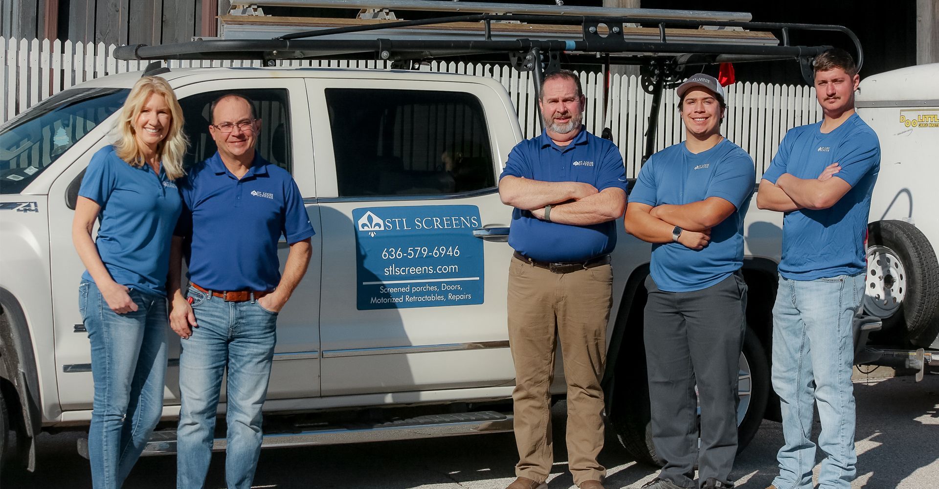 Five people in blue shirts pose beside a work truck with arms crossed, smiling.