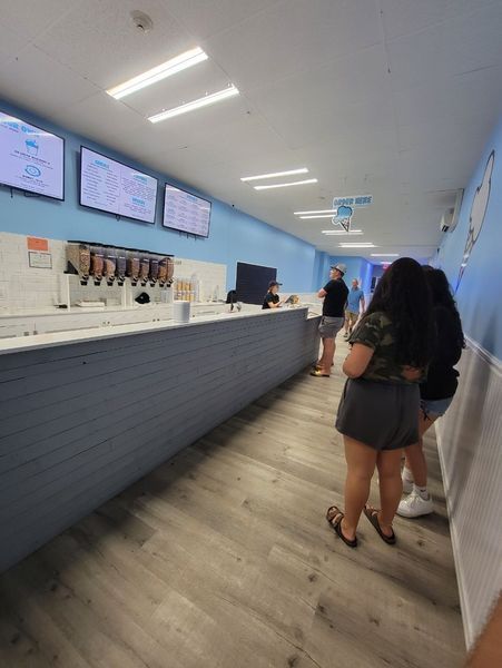 Inside a blue-themed shop, customers queue at the counter. A long, gray counter is lined with beverage dispensers.