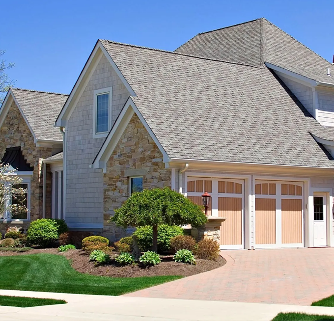 Beige house with stone accents, tan garage doors, and landscaped yard under a blue sky.