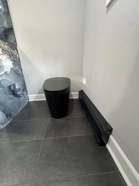 Black toilet in a corner of a bathroom with gray tile floor and walls, next to a black speaker.