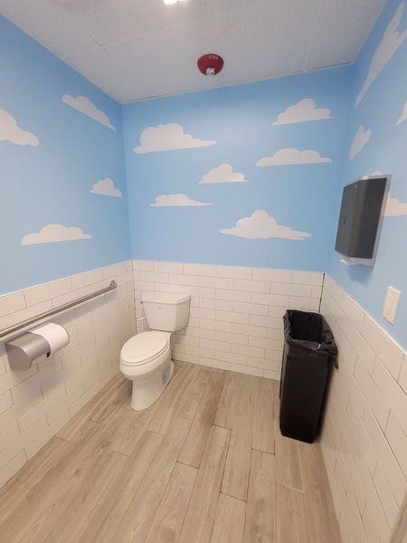 Restroom with a blue sky and cloud mural, white tiled walls, and a toilet.