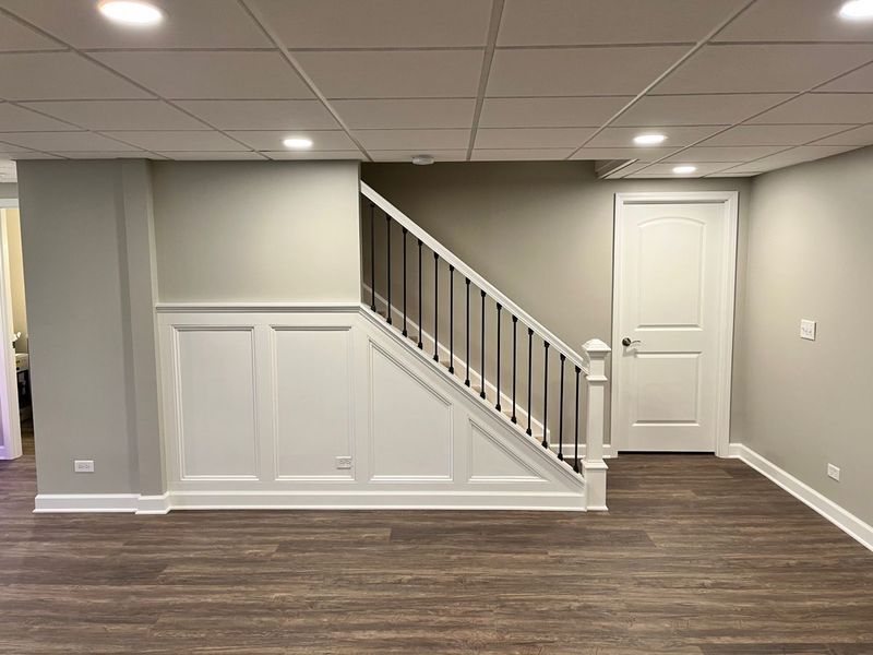 Basement with stairs, wainscoting, a white door, and wood-look flooring; neutral paint colors.