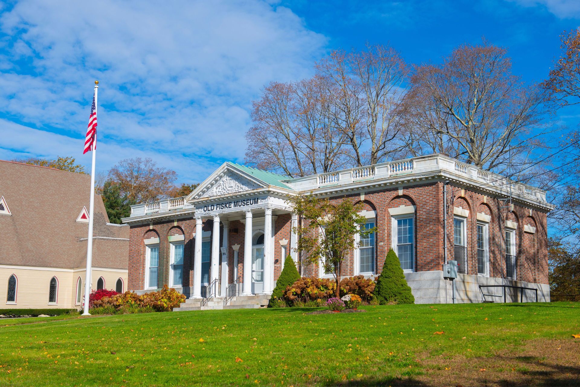 White-columned brick civic building with an American flag, set on a grassy lawn under a blue sky