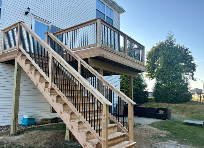 Wooden deck with stairs, black railing, attached to a two-story white house with blue sky background.