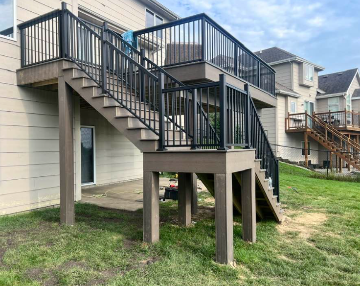 Deck with black railings and stairs in a grassy backyard, adjacent to a beige house and other houses.