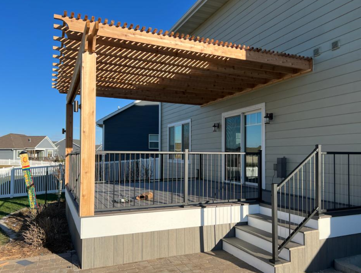 Wooden pergola on a deck attached to a house with sliding doors. Gray railing and steps lead down.