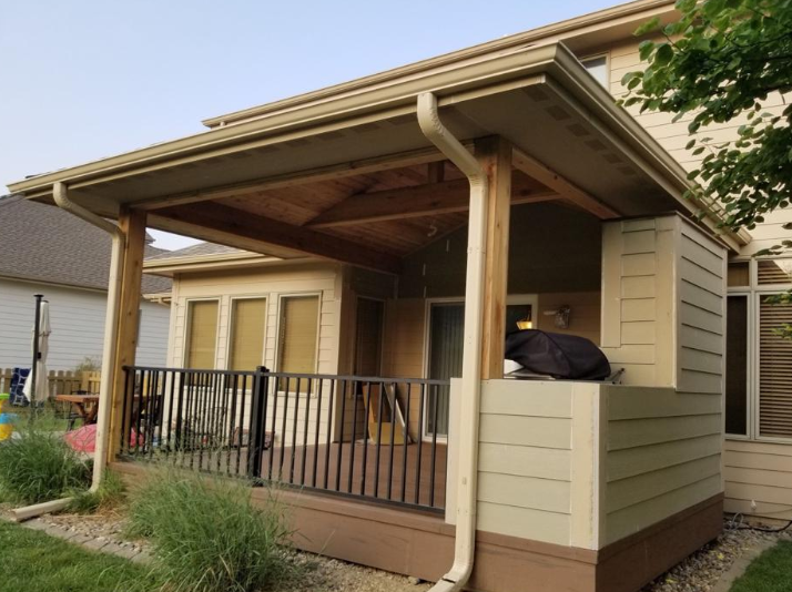 Covered patio with brown railing, tan siding, and light-colored gutters.