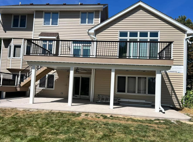 Back view of a two-story beige house with a deck and patio. The deck has black railings, and the patio is concrete.