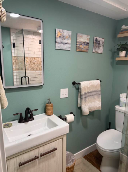 Bathroom with green walls, white sink, toilet, mirror, and artwork. A towel hangs on a black bar.