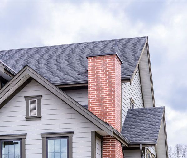 A brick chimney rises from a gray-roofed house with light-gray siding.