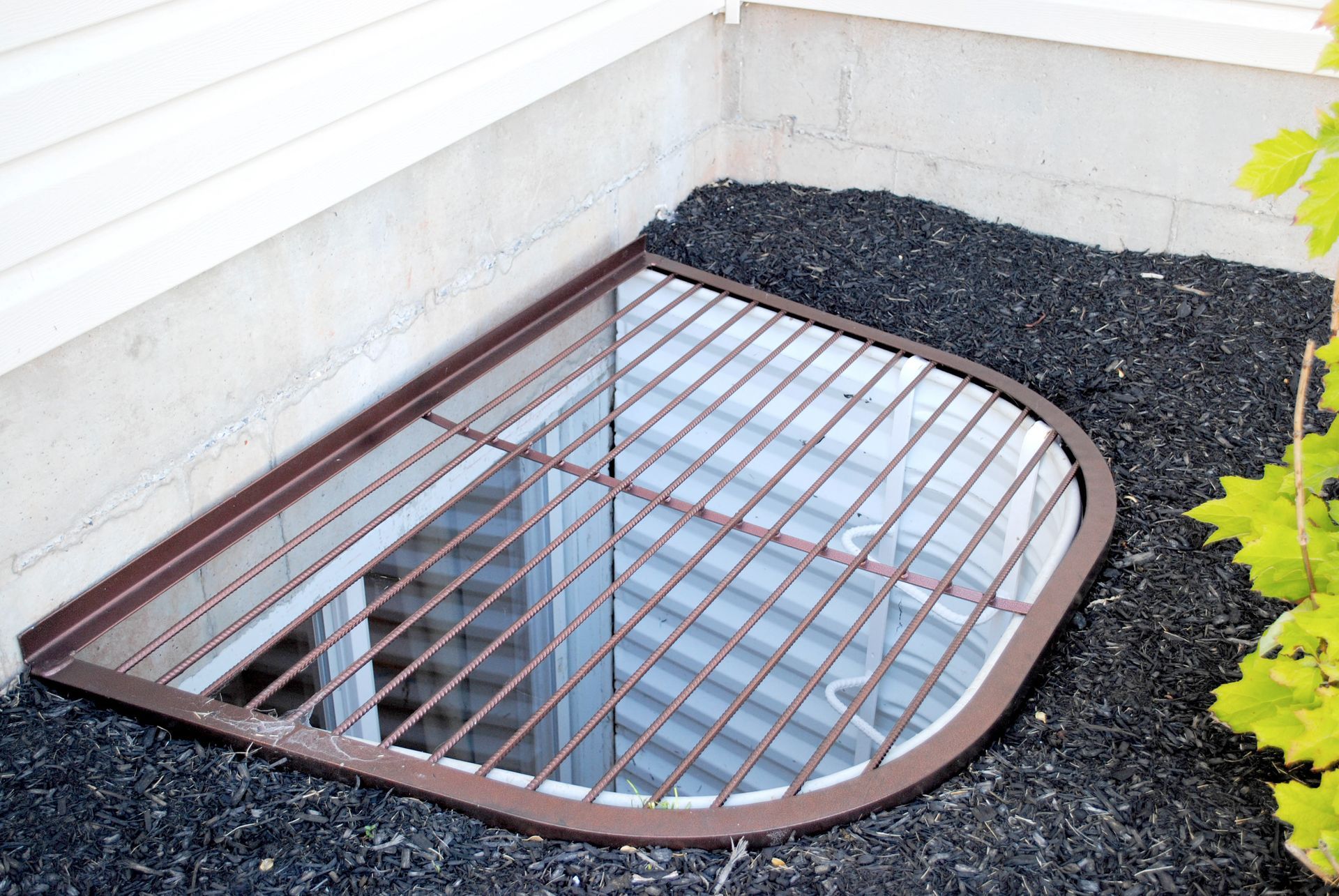 Brown metal window well with grated cover, surrounded by black mulch, next to a building.
