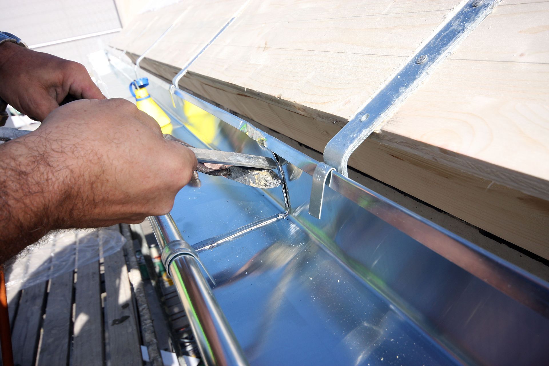 Hands using pliers to repair a metal gutter on a wooden structure.