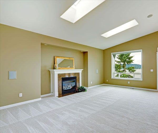 Empty living room with fireplace, window, skylights, beige walls and carpet.