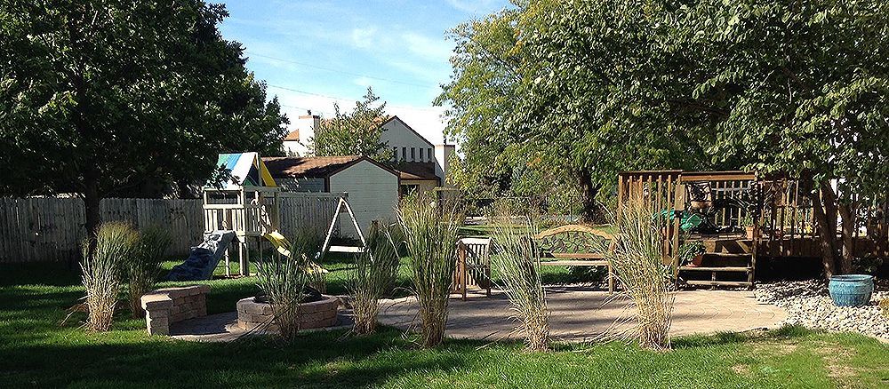 A backyard playground with a swing set, wooden play structure, bench, and greenery under a blue sky.