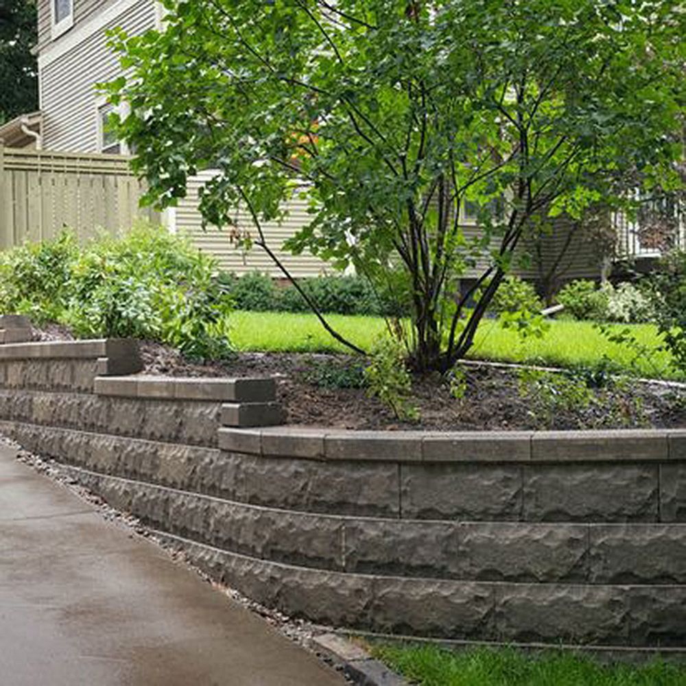 Gray stone retaining wall with greenery and a tree, next to a driveway.