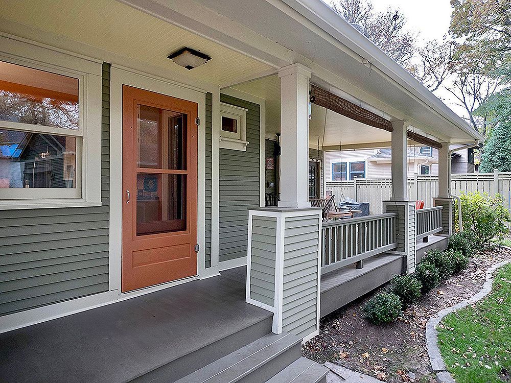 Green house exterior with brown door, porch, gray steps, and small bushes along the lawn.