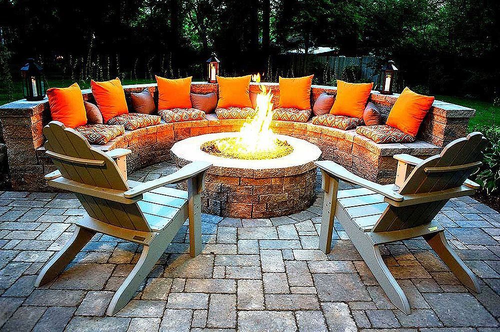 Outdoor fire pit with seating: two chairs face the fire, a curved bench with orange cushions surrounds the pit.