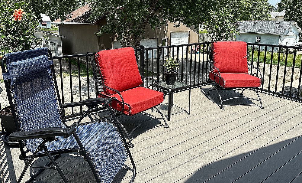 Deck with three chairs; two red cushions, one blue, all facing toward a cityscape with houses and a garage.