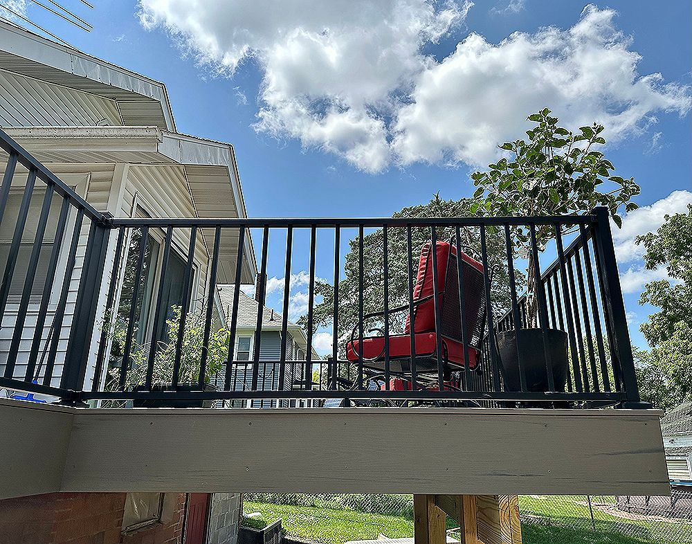 Black railing on a deck with red chair and potted plant against a cloudy blue sky.