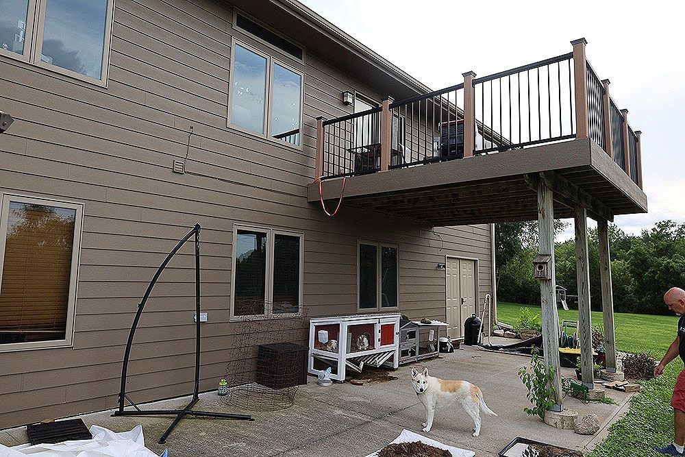 Two-story house with a wooden deck. A dog stands on a patio. Person in shorts near plants.