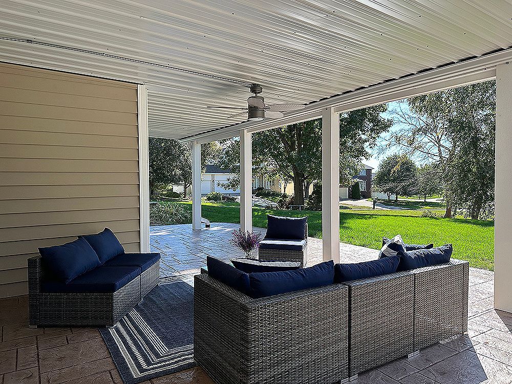 Covered patio with wicker furniture, rug, and ceiling fan, overlooking a green yard and houses.