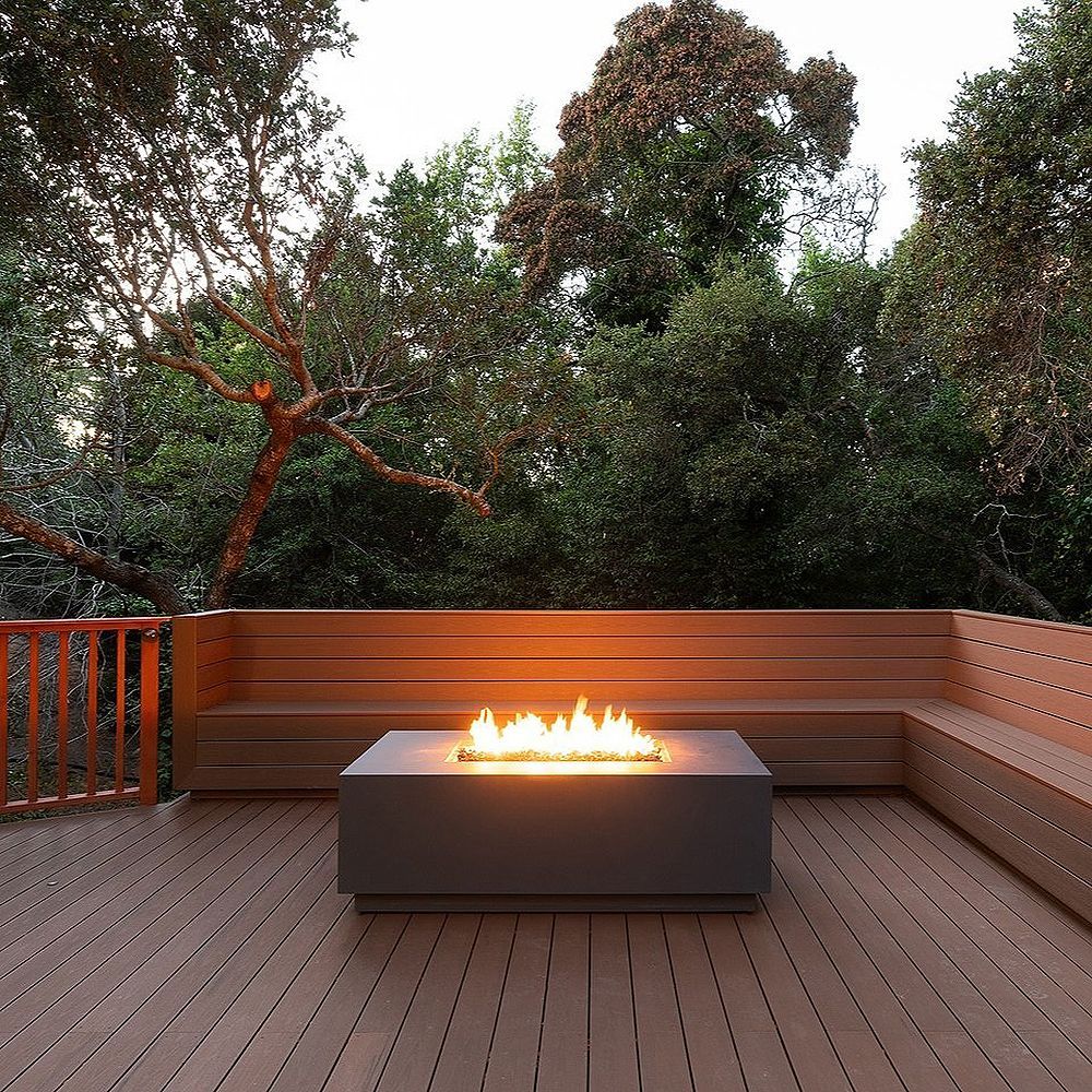 Modern outdoor deck with fire pit and built-in benches, surrounded by trees.