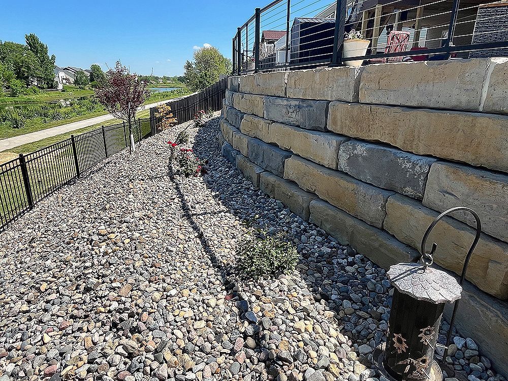Stone retaining wall and gravel path alongside a black metal fence, on a sunny day.