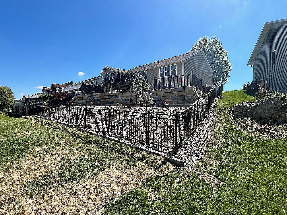 A house on a hillside with a stone retaining wall and black metal fence under a blue sky.