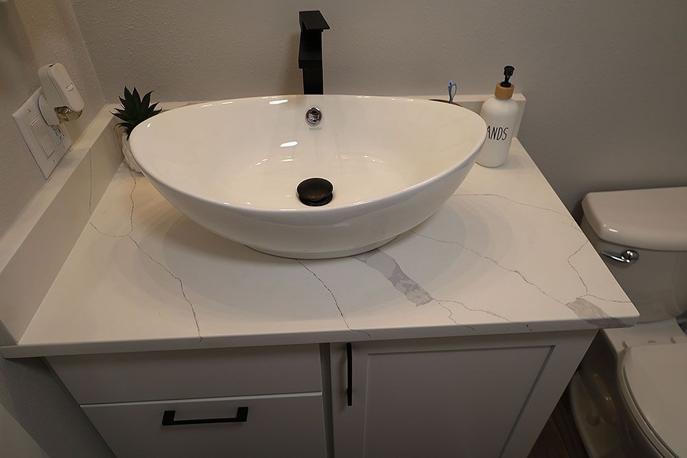 White oval vessel sink on a white countertop with a black faucet. Light gray cabinet below.