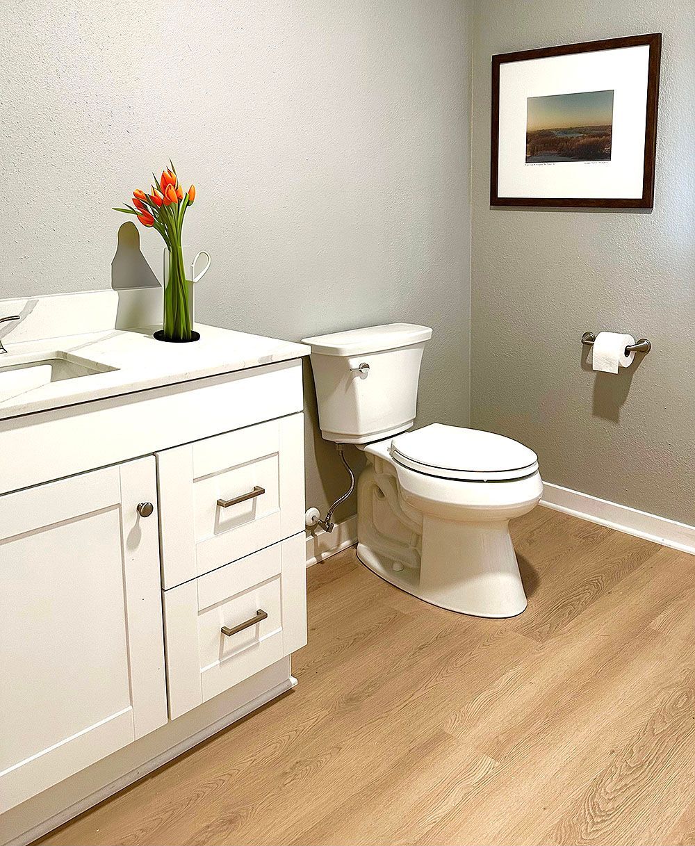 White bathroom with vanity, toilet, floral arrangement, and framed artwork. Light wood-look flooring and silver textured walls.