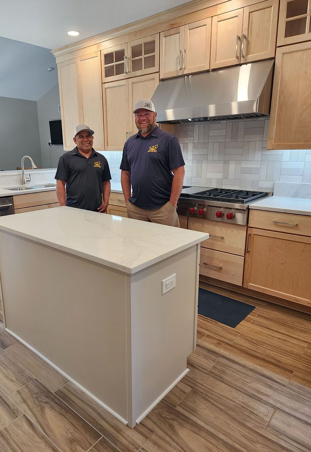 Two men in a remodeled kitchen with light wood cabinets, an island, and a stainless steel range hood.