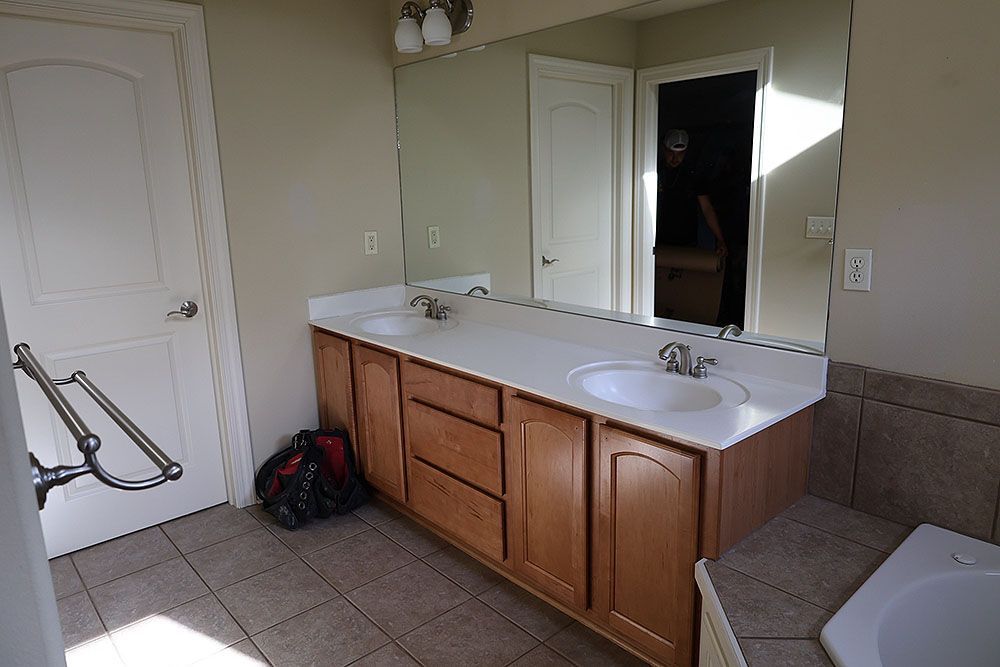 Bathroom with double vanity, large mirror, and open door. Brown cabinets, white counter and sinks, beige walls, and tile floor.