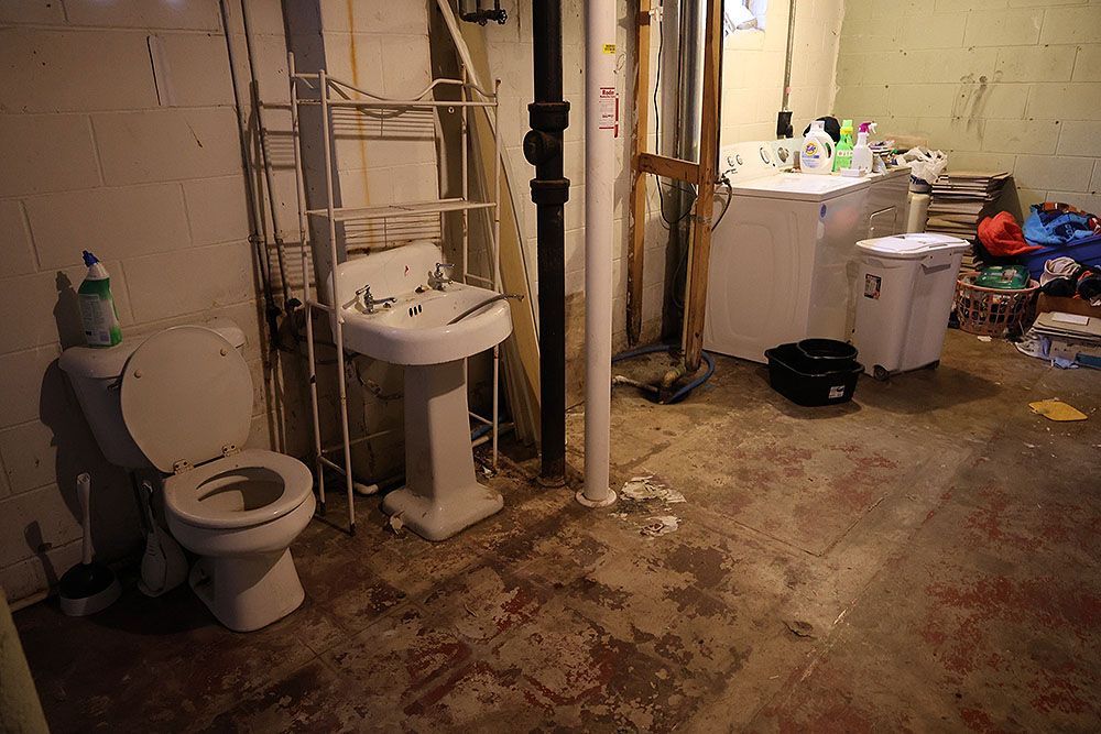 Basement bathroom with a toilet, sink, and washing machine. Floor is stained, items scattered.