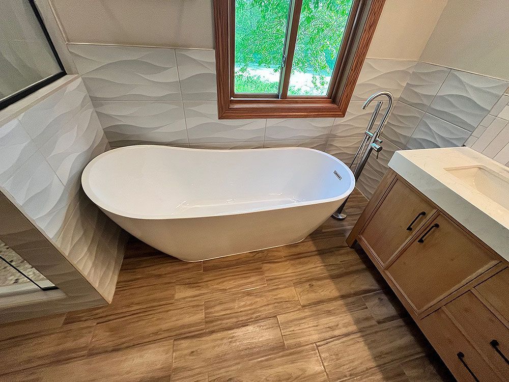Bathroom with a white freestanding tub, wood-look tile floor, light wood vanity, and textured white wall tiles.