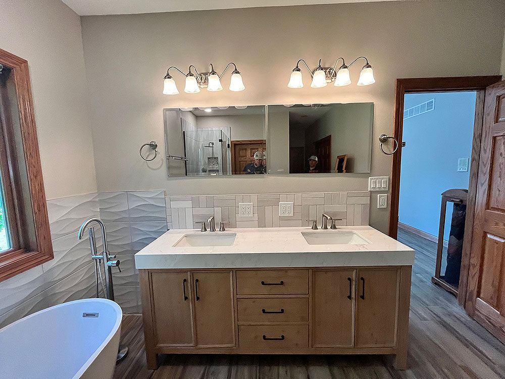 Bathroom with light wood vanity, dual sinks, large mirrors, and a freestanding tub.