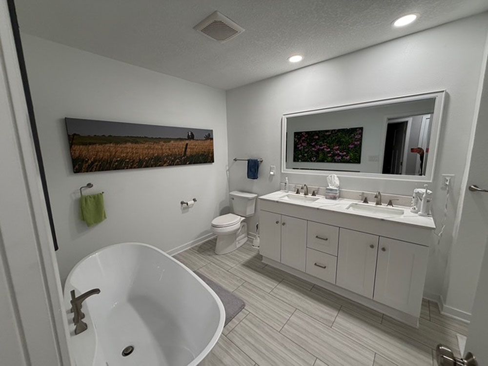 Bathroom with white tub, double vanity, artwork, and grey tile floor.
