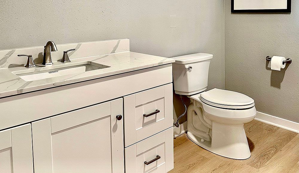 White bathroom with vanity, toilet, and paper holder against a textured gray wall; wood-look floor.