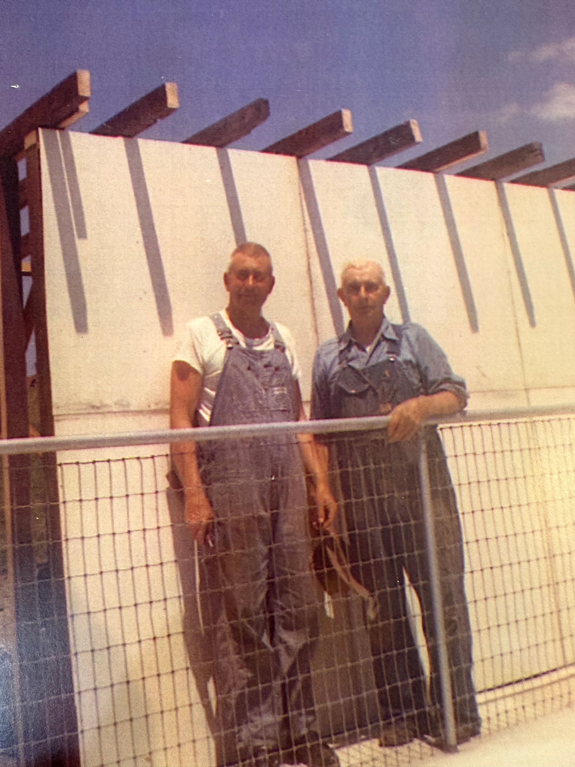 Two people in work overalls stand side-by-side in front of a white wall with overhead beams and a wire fence.