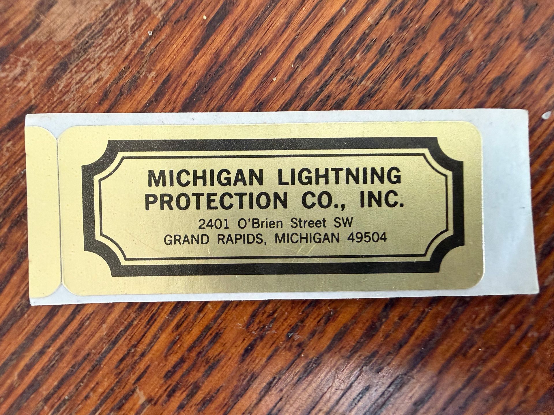 A gold sticker on a white backing, reading MICHIGAN LIGHTNING PROTECTION CO., INC. - 2401 O'Brien Street SW Grand Rapids.