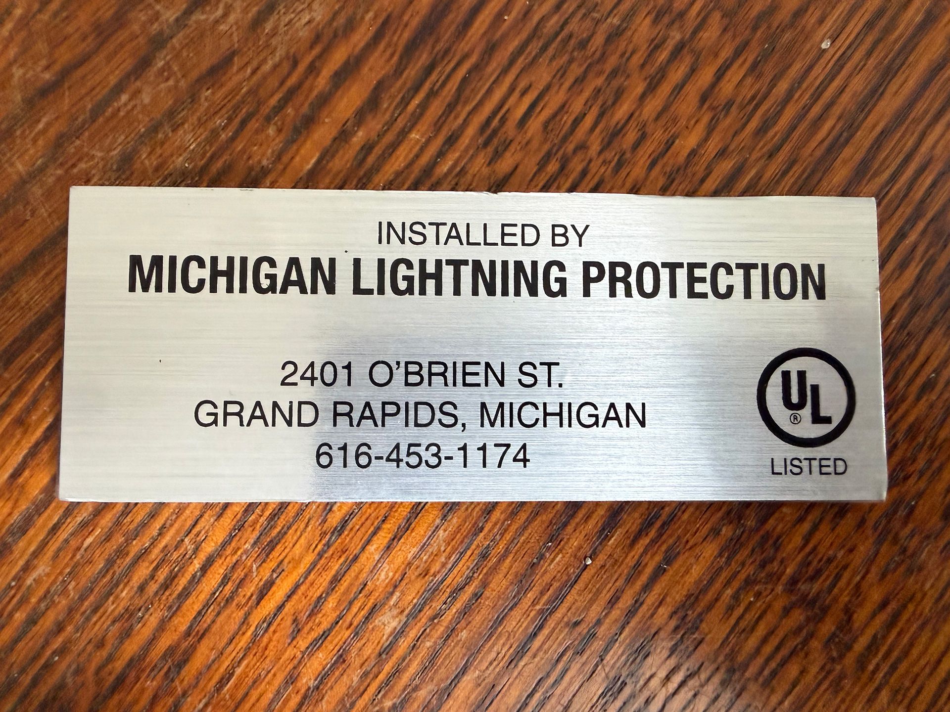 A silver metal tag for Michigan Lightning Protection in Grand Rapids, MI, with contact details and a UL Listed logo.