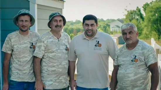 Four men pose on a porch. Three in camouflage shirts, one in a tan polo. Green trees in the background.