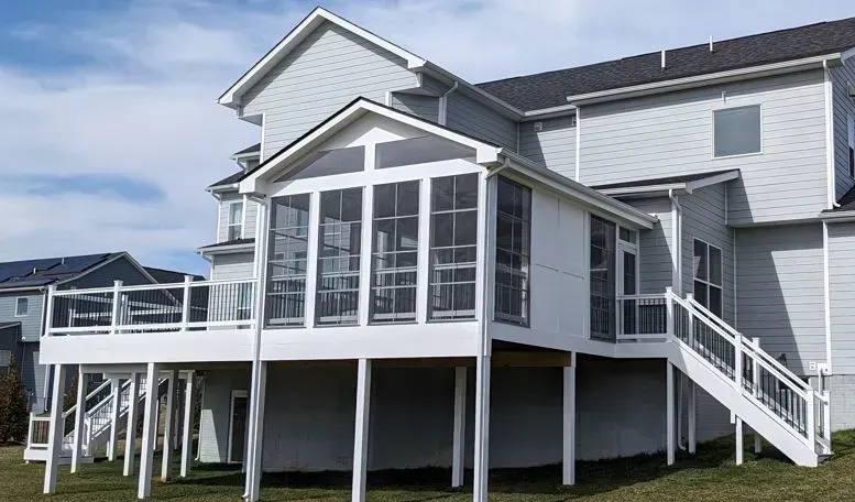 White screened porch and deck attached to a gray house, white railings, blue sky.