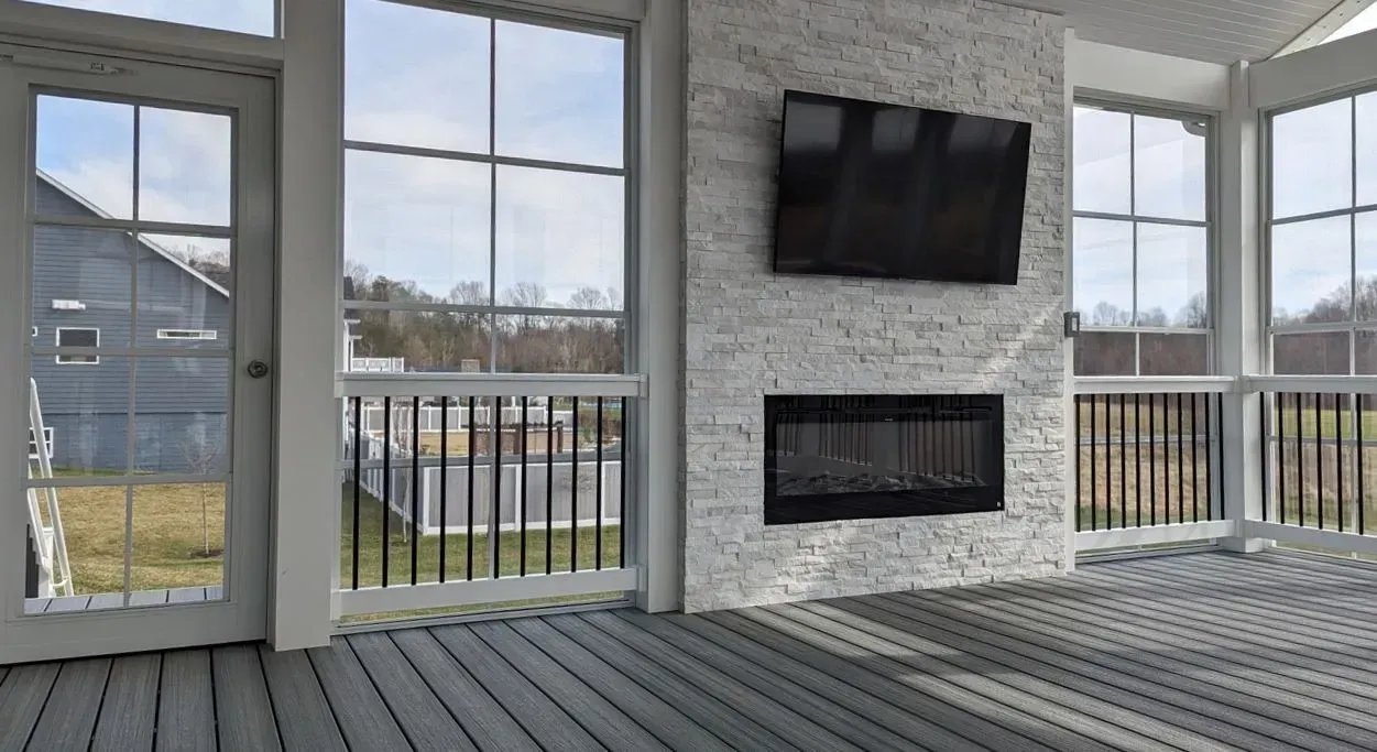 Sunroom with gray flooring, white brick wall, fireplace, TV, and large windows.